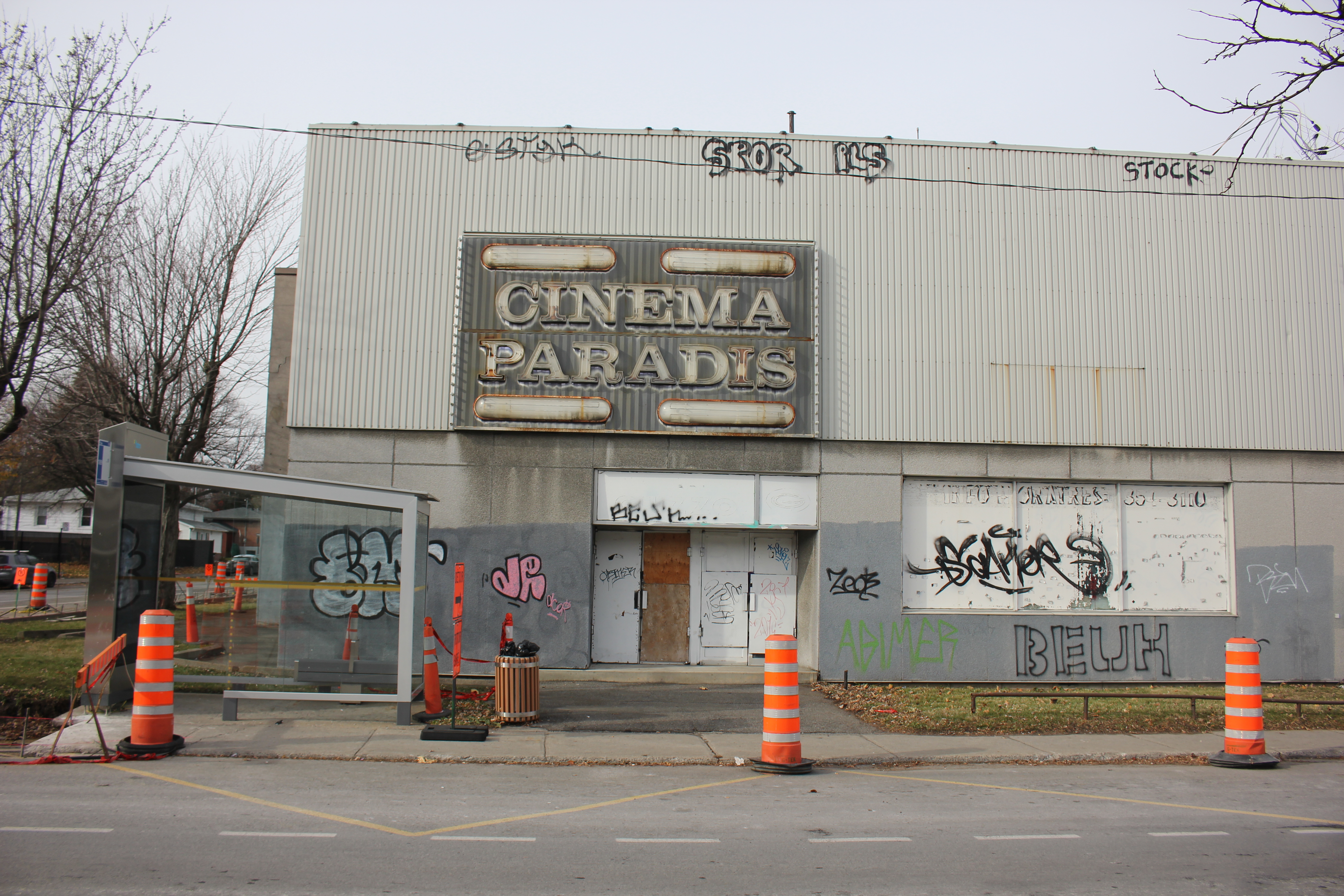 La façade du bâtiment délabré de l'ancien cinéma Paradis, montrant des portes et fenêtres barricadées et des graffitis. Des cônes oranges sont placés devant le bâtiment.