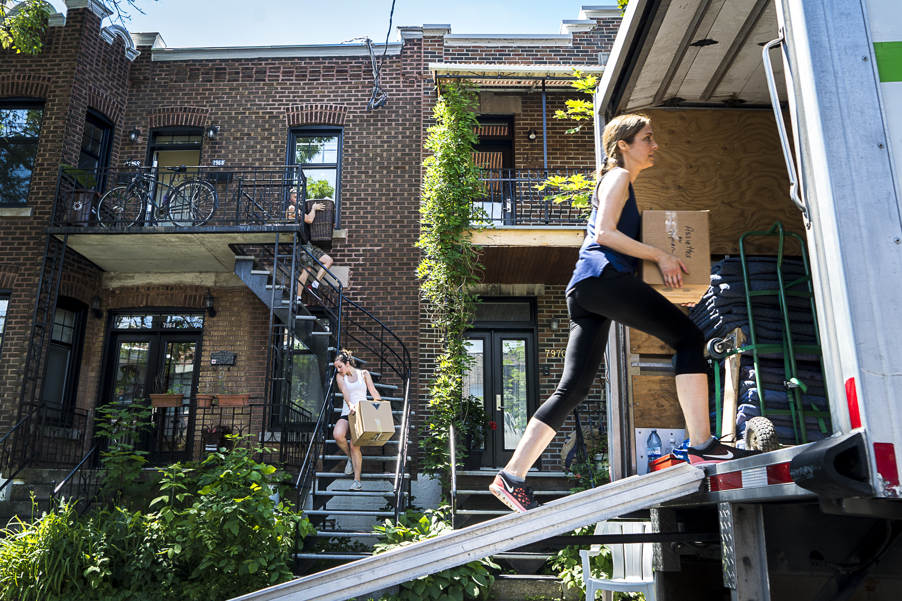 Une femme transporte une boîte dans un camion lors de la journée du déménagement du 1er juillet. En arrière plan, deux autres personnes descendent des escaliers avec des boîtes.
