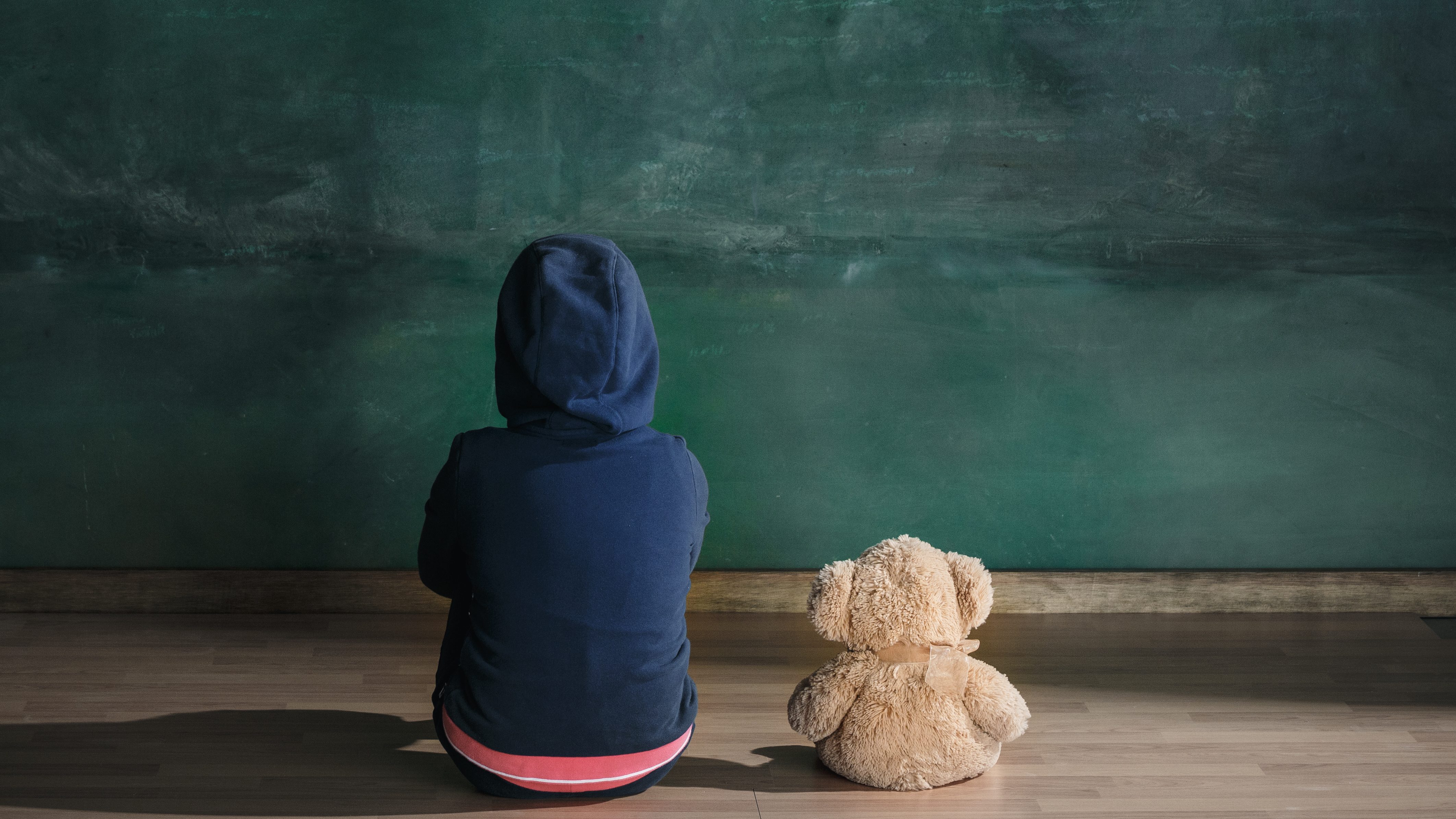 Un enfant d'un milieu défavorisé regarde un tableau vert, de dos, à côté d'un nounours.