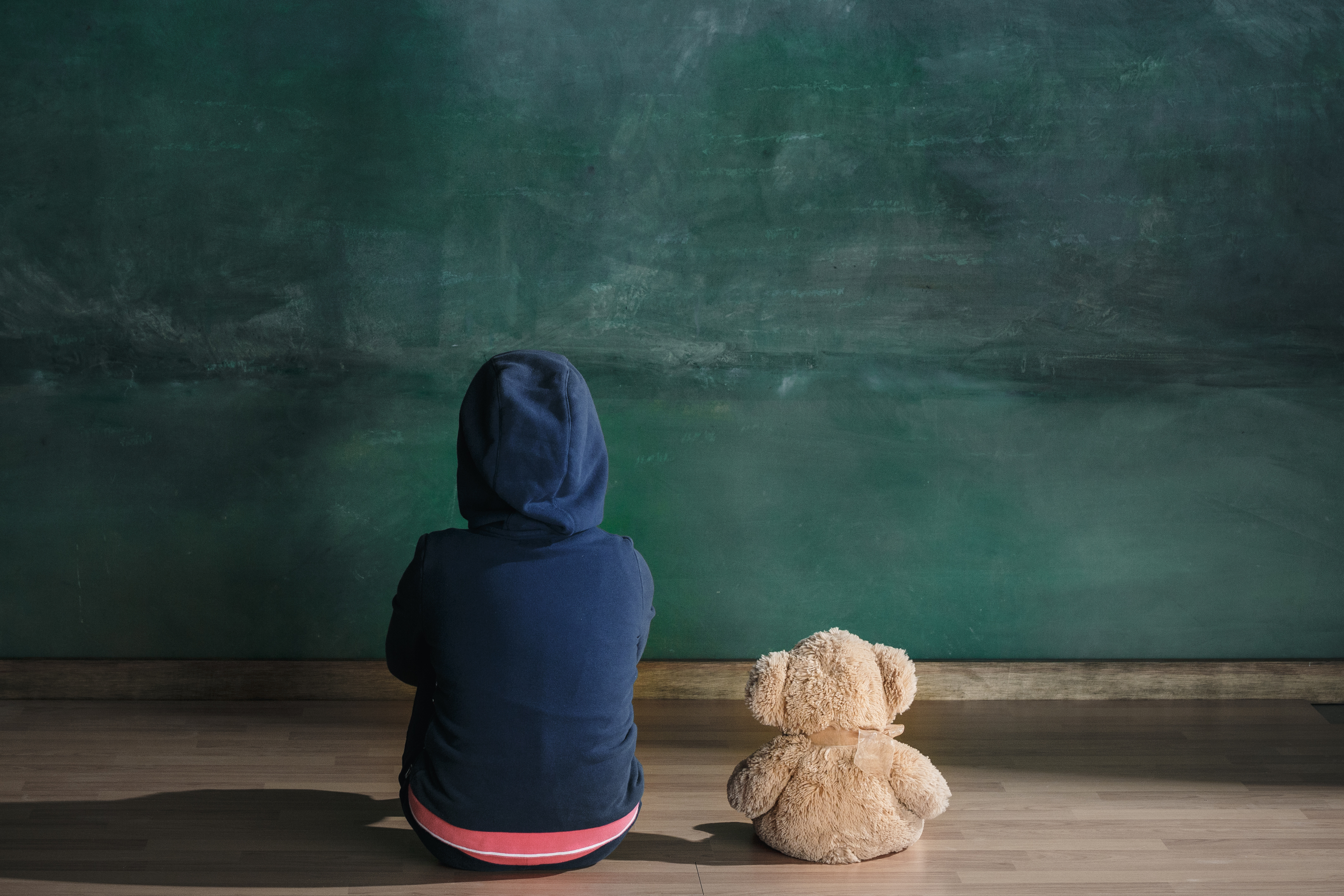 Un enfant d'un milieu défavorisé regarde un tableau vert, de dos, à côté d'un nounours.