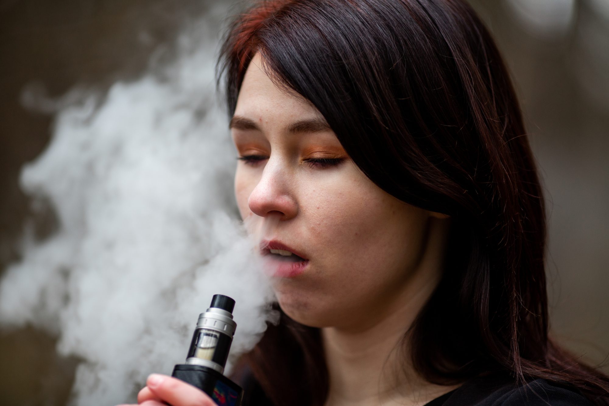 Vape teenager. Young pretty caucasian brunette girl smoking an electronic cigarette on the street in the spring. Deadly bad habit.