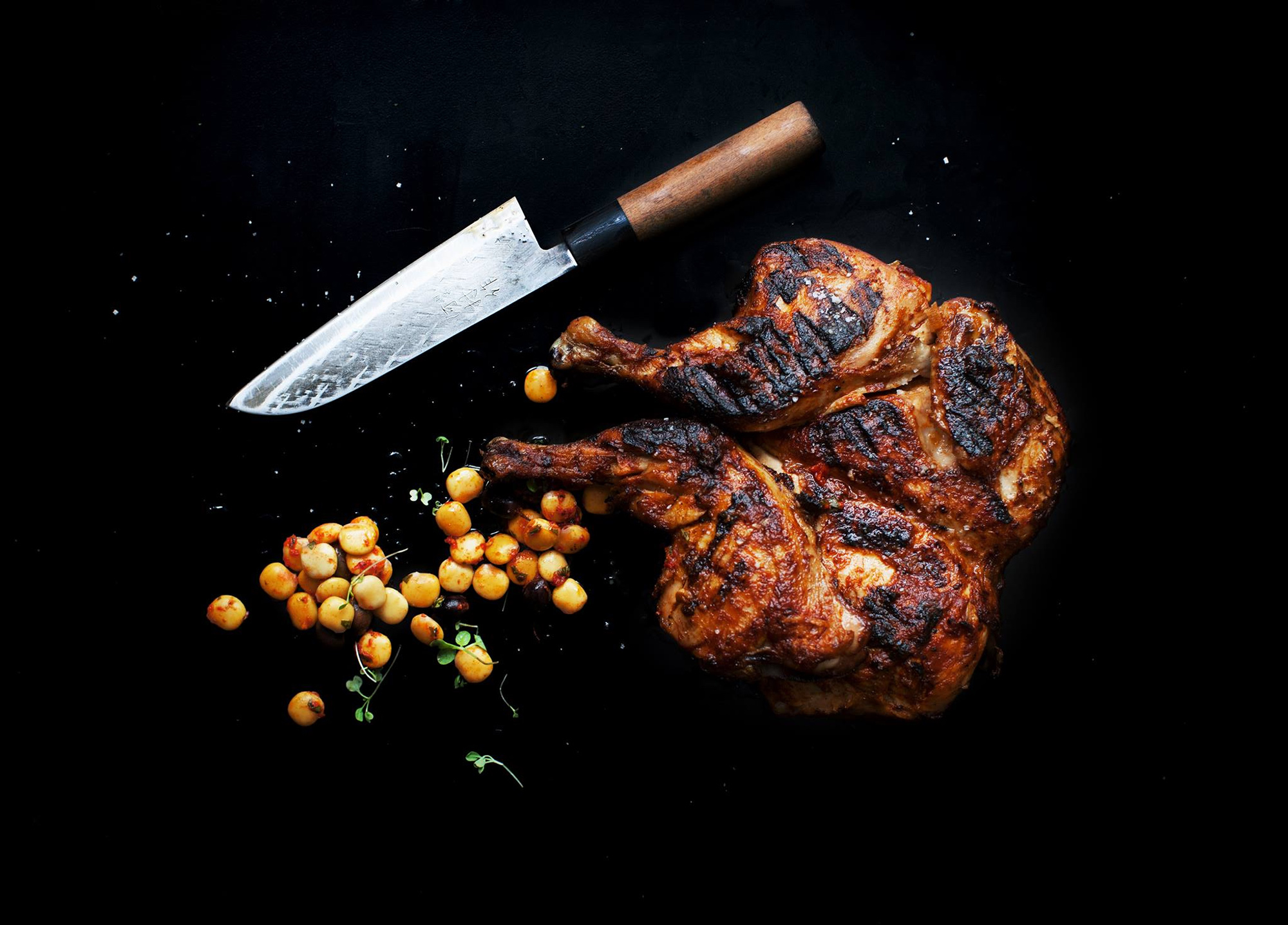 Poulet grillé en crapaudine sur fond noir. Couteau japonais et grains de maïs.