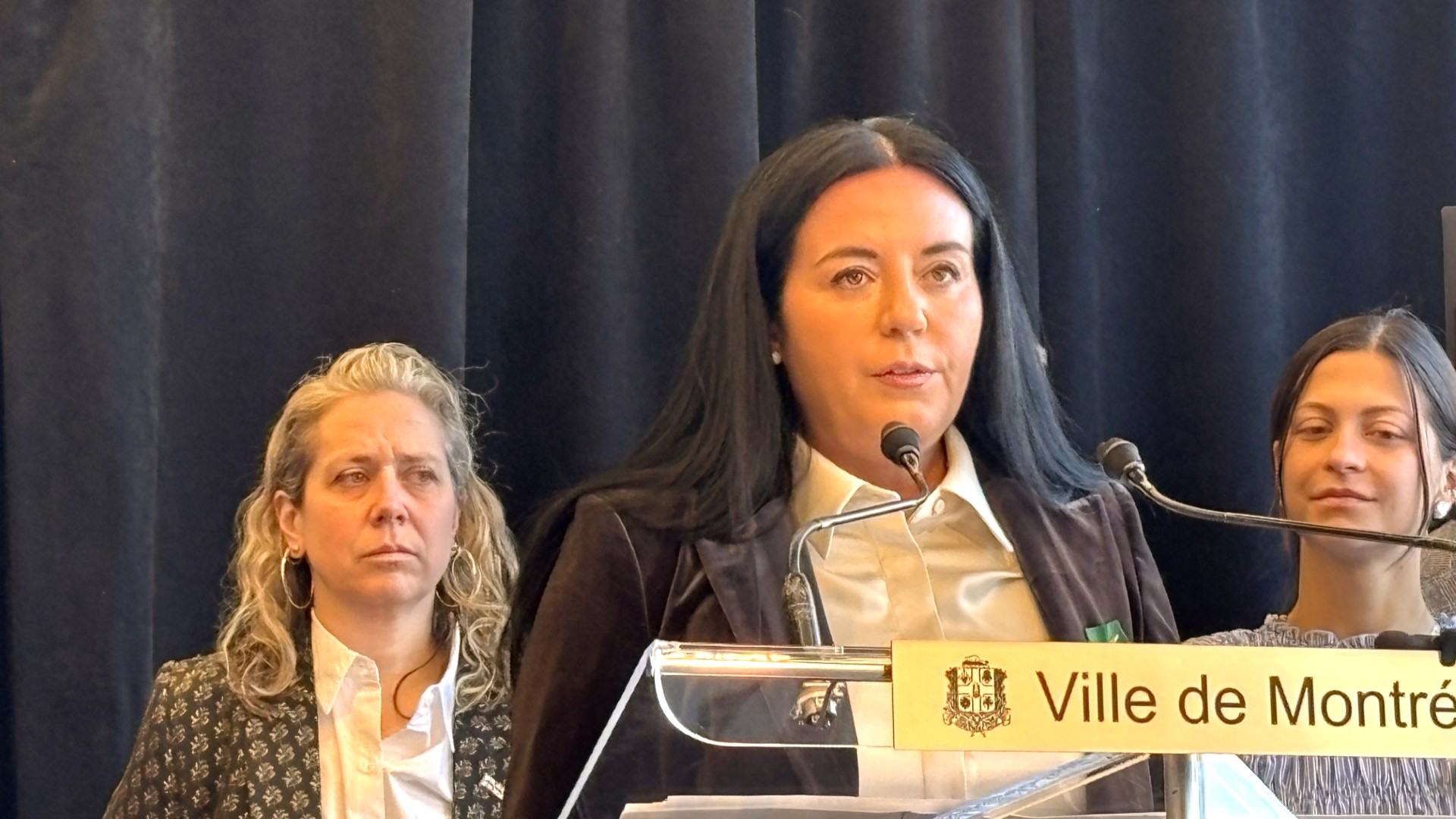 Soraya Martinez Ferrada devant un lutrin. Derrière elle se trouvent les conseillères Caroline Braun et Mercedez Plante.