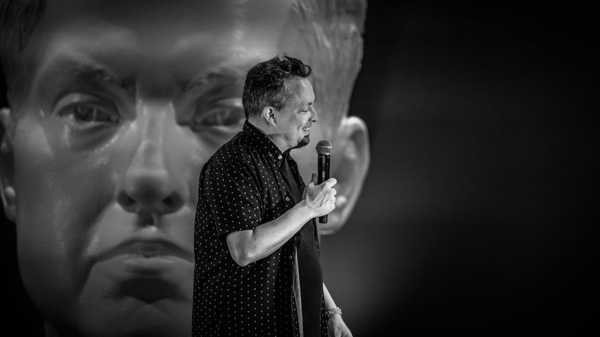 Image en noir et blanc de Mike Ward sur scène devant un buste géant de lui-même.