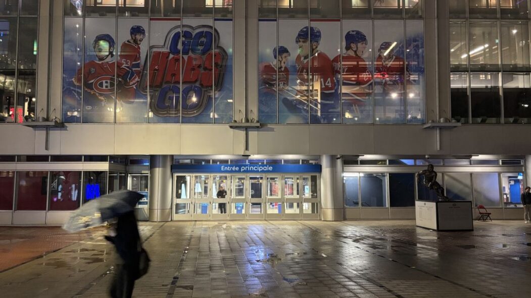 Une personne marche sous la pluie devant l'entrée principale du Centre Bell, la nuit.